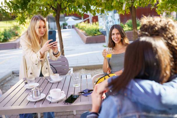 Jeune fille blonde prenant une photo avec un smartphone de ses amis assis à une table de bar. - Photo, image