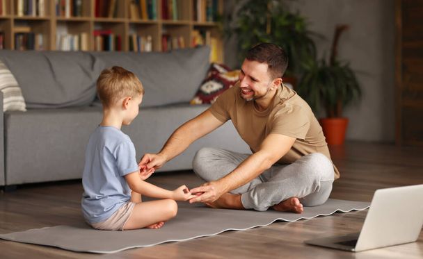 Genç pozitif baba, Lotus 'ta birlikte oturma odasında poz verirken ve dizüstü bilgisayardan yoga dersleri izlerken nasıl meditasyon yapılacağını gösteriyor. - Fotoğraf, Görsel