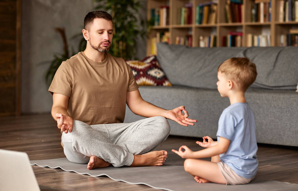 Семья практикует йогу онлайн с ноутбуком. Молодой человек отец и маленький ребенок сын в повседневной одежде медитируют вместе, сидя в позе лотоса на полу перед ноутбуком в гостиной - Фото, изображение