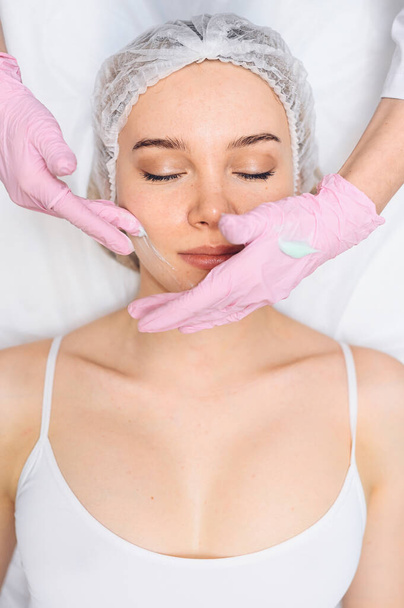 Beautiful happy woman receiving spa treatment. Cosmetologist in beauty salon with a pink glove applies a moisturizing cream mask. Facial beauty. Perfect fresh clean skin. Youth and skin care concept  - Photo, Image