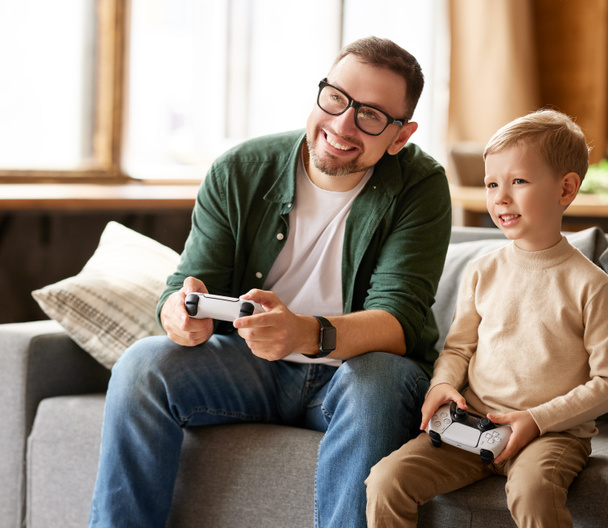 Real competition. Happy family, man father and excited boy son playing video console games together while spending leisure time together at home, sitting on couch in living room at home - Photo, Image