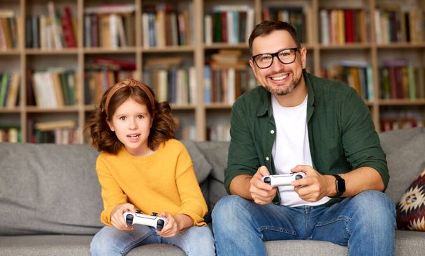 Prawdziwa konkurencja. Szczęśliwa rodzina, mężczyzna ojciec i podekscytowany córka gra w gry wideo konsoli razem podczas spędzania wolnego czasu razem w domu, siedzi na kanapie w salonie w domu - Zdjęcie, obraz