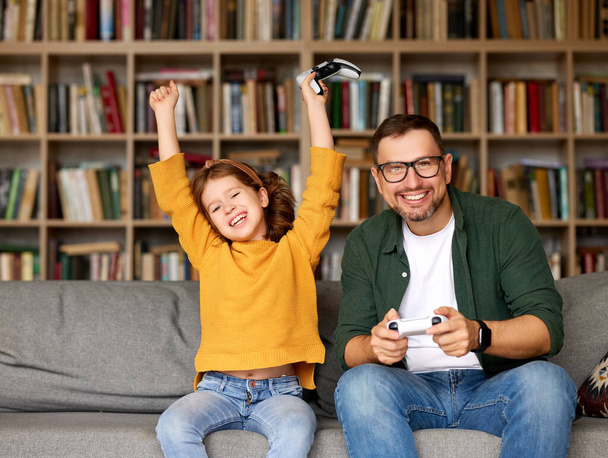 Young happy man father and excited daughter celebrating victory while playing video console games together, playful dad spending time with child while relaxing on couch in living room at home - Foto, Bild