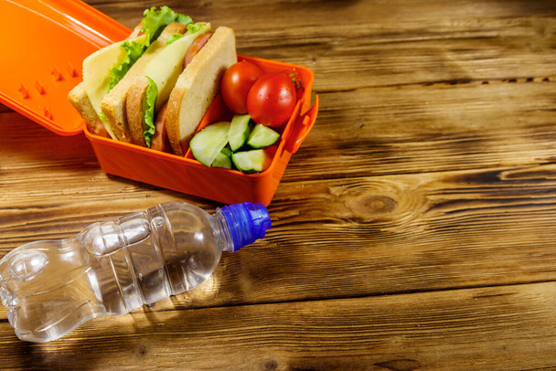 Bottle of water and lunch box with sandwiches and fresh vegetables on a wooden table - Photo, Image
