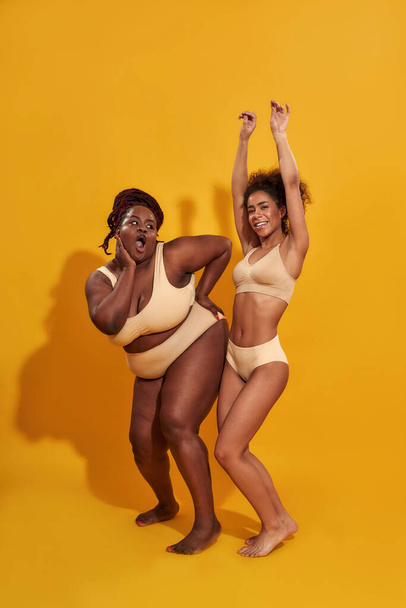 Full length shot of joyful african american women in beige underwear with different body size looking playful while posing isolated over yellow background - Fotoğraf, Görsel