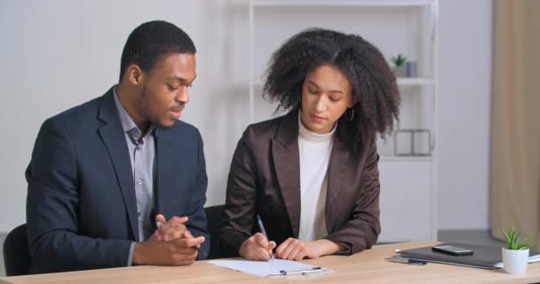 Dwóch afro amerykańskich partnerów biznesowych para etnicznych osób siedzących przy stole czytając dokument kobieta doradca prawnika konsultant pokazujący pracownikowi umowy o współpracy papierowej oferty do podpisania - Materiał filmowy, wideo