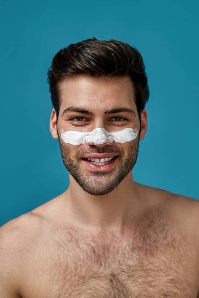 Good looking shirtless guy with clean skin and white mask applied on nose for cleansing pores smiling at camera isolated over blue background - Fotografie, Obrázek