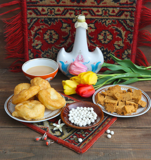 Cazaque ainda vida no costume nacional de usar brincos "Syrga salu", com comida nacional: baursaks, irimshik, kurt, koumiss e chá em um fundo de madeira - Foto, Imagem