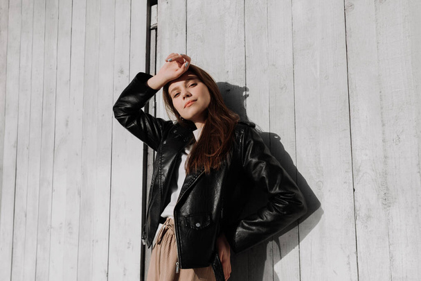 Улыбающаяся молодая женщина в черной кожаной куртке поднимает руку, стоя перед белой деревянной стеной. Веселимся на улице. Городской стиль и уличная мода. Девушка в повседневной одежде смотрит в камеру - Фото, изображение