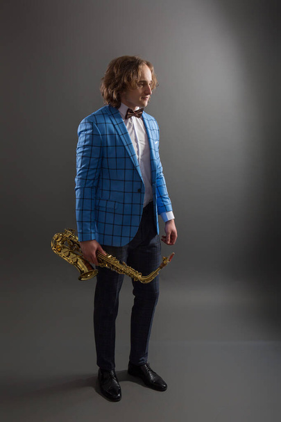Полнометражный студийный портрет молодого музыканта в ярко-голубой клетчатой куртке с саксофоном. - Фото, изображение