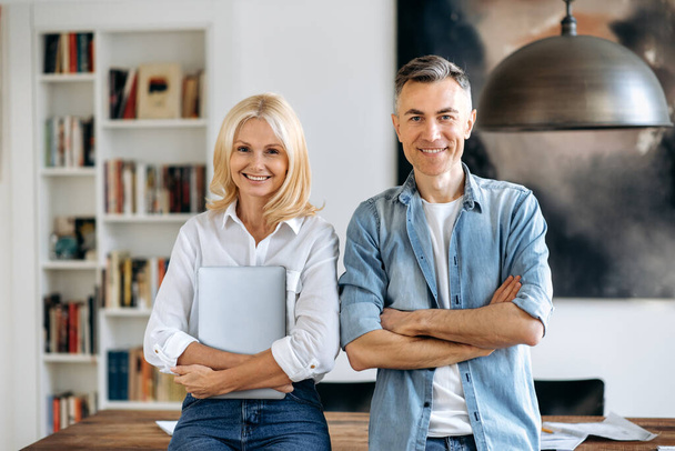 Portrait of two mature happy caucasian intelligent business partners in stylish wear, stand near desk, arms crossed, look at camera, smiling friendly. Charming business lady, attractive business man - Photo, Image