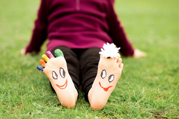 Mała dziewczynka siedzi na trawie z malowanymi stopami z zabawnymi komicznymi twarzami - Zdjęcie, obraz