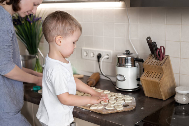 ポートレートかわいい小さな幼児白人の男の子家庭のキッチンで若い大人の母親と甘いおいしいクッキーを調理します。ママの小さな息子は甘いペストリー手作りデザートを焼く。親子で一緒に活動 - 写真・画像