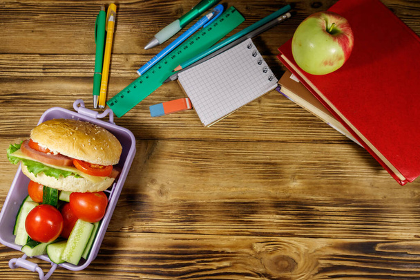 De vuelta al concepto escolar. Material escolar, libros, manzana y lonchera con hamburguesas y verduras frescas sobre una mesa de madera. Vista superior - Foto, Imagen