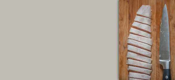 Banner con pescado medio congelado en una tabla de madera cortada en rodajas finas por un cuchillo grande en un fondo sólido con espacio de copia para texto, detalles, primer plano. Concepto de cocina casera y comida saludable. - Foto, Imagen