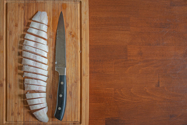 Полузамороженная рыба на деревянной доске, разрезанной на мелкие кусочки большим ножом, детали, крупным планом, с копировальным местом в виде деревянной столовой тарелки. Концепция домашней кухни и здорового питания - Фото, изображение
