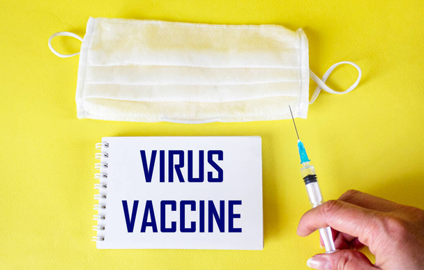 Auf gelbem Hintergrund eine Medina-Maske und ein Notizbuch mit dem Text VIRUS VACCINE. Weibliche Hand mit Impfstoff - Foto, Bild