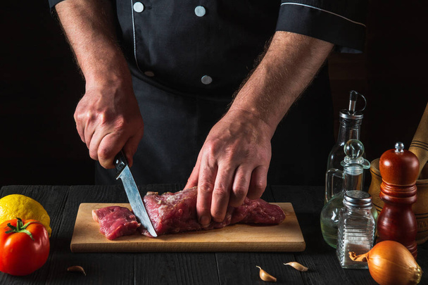 Шеф-кухар, який ріже яловиче м'ясо з ножем на кухні, готує їжу. Овочі та спеції на кухонному столі для приготування смачного обіду
 - Фото, зображення