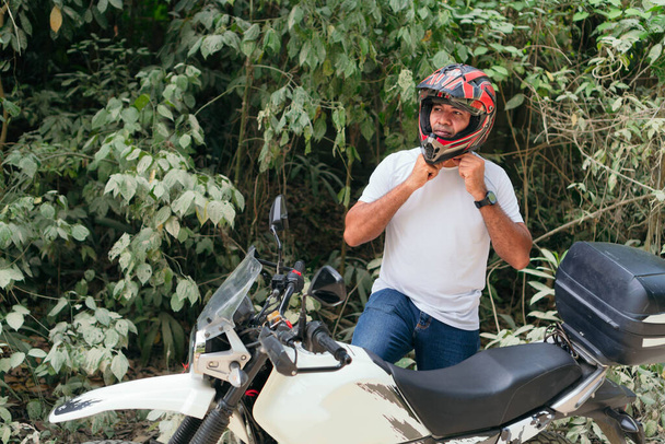 Młody mężczyzna w białej koszuli i dżinsach przypięty do kasku na motocyklu na świeżym powietrzu. - Zdjęcie, obraz
