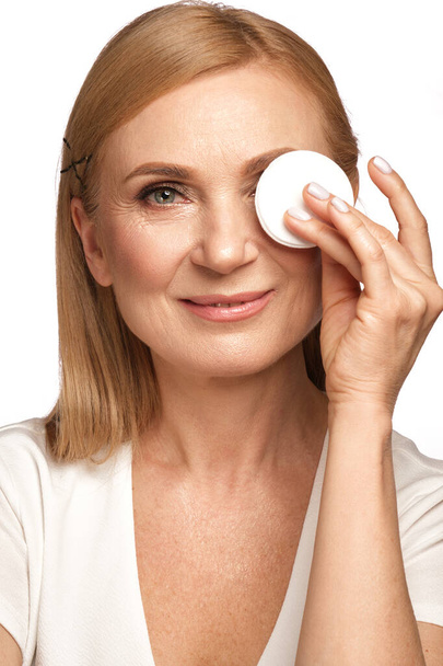 Portrait of a beautiful elderly woman in a white shirt removing makeup with a cleanser and a cotton pad. - Foto, Bild