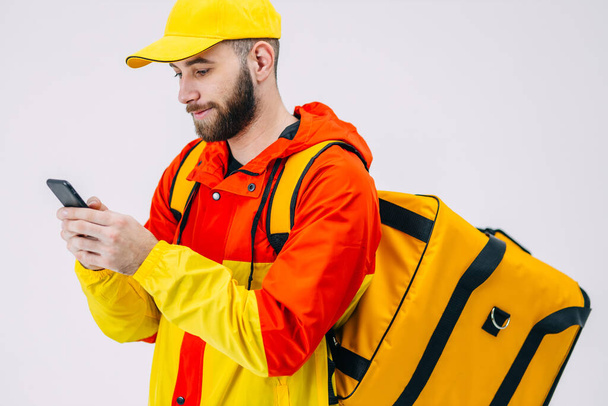 Elinde bir sırt çantası ve akıllı telefonu olan bir kurye. Sarı ceketli bir kurye emir beklerken sosyal medyayı kontrol ediyor.. - Fotoğraf, Görsel