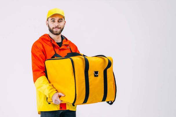Бородатый улыбающийся мужчина из службы доставки держит рюкзак в красно-желтой форме и кепку в студии на белом фоне. Доставка еды. - Фото, изображение