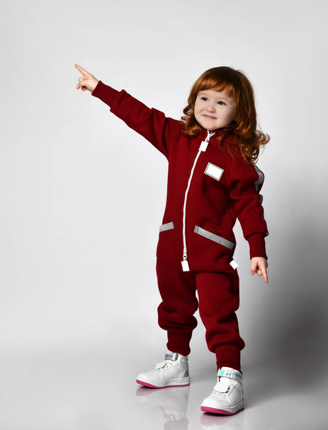 Joyful little red-haired toddler girl in warm sports overalls side view portrait - 写真・画像