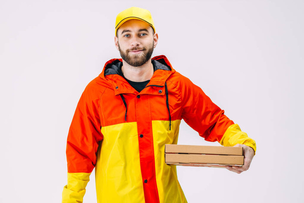 Primer plano de guapo joven barbudo repartidor sosteniendo cajas de pizza, dando orden en uniforme rojo y amarillo y gorra. - Foto, Imagen