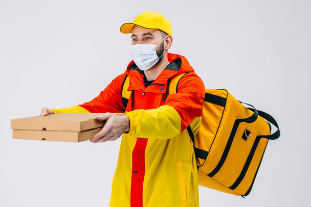 Ein glücklicher Kurier mit medizinischer Maske hält eine Pizza in Kartons und präsentiert sie einem Kunden. Bekleidet mit einer rot-gelben Uniform und einer Kühltasche auf dem Rücken. Kurierlieferung von Lebensmitteln nach Hause. - Foto, Bild