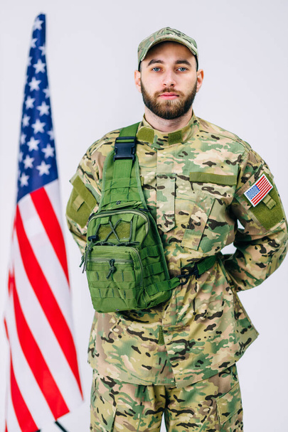 Een jonge soldaat met baard in uniform, met zijn handen achter zijn rug, met een ernstig gezicht, vlakbij de vlag. - Foto, afbeelding