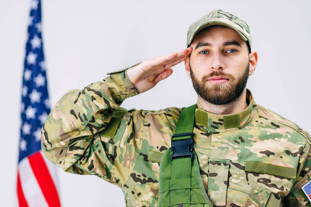 der gut aussehende Soldat in Militäruniform salutiert und blickt in die Kamera neben der amerikanischen Flagge im Studio auf weißem Hintergrund - Foto, Bild