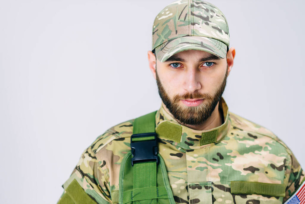 Vista de cerca de un soldado serio en uniforme militar mirando a la cámara cerca de la bandera americana en el estudio sobre un fondo blanco - Foto, imagen