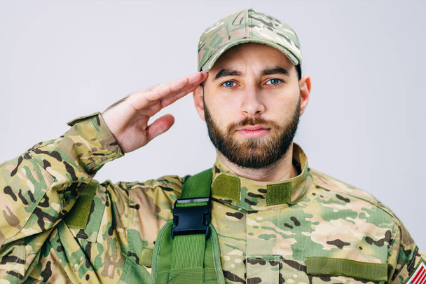 Portrét vojáka americké armády v maskovací uniformě, jak salutuje, dívá se do kamery. Voják si drží ruku u hlavy jako projev úcty.. - Fotografie, Obrázek