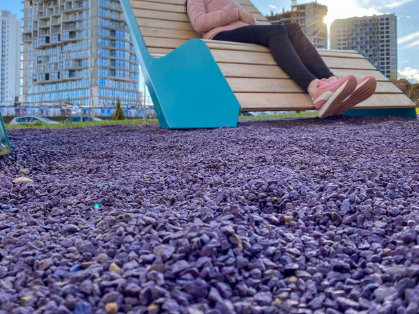 A woman in beautiful sportswear with pink sneakers on her feet sits on a fashionable new bench lounger in the park with purple pebbles for landscape design. - Photo, Image