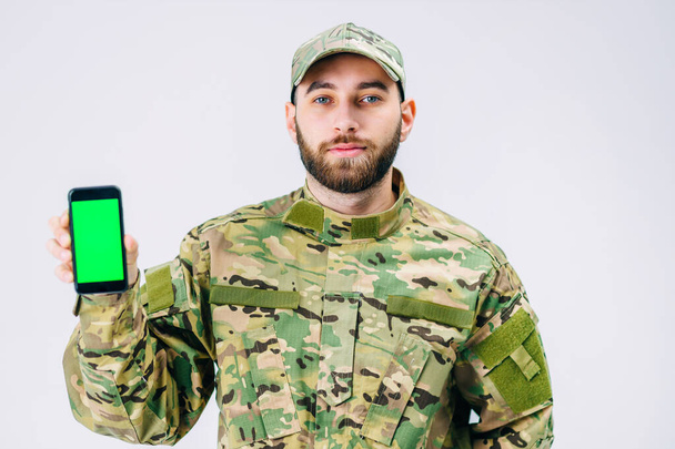 Atraktivní voják ve vojenské uniformě ukazuje svůj smartphone zelené obrazovce chroma klíč s úsměvem a dívá se do kamery ve studiu na bílém pozadí - Fotografie, Obrázek