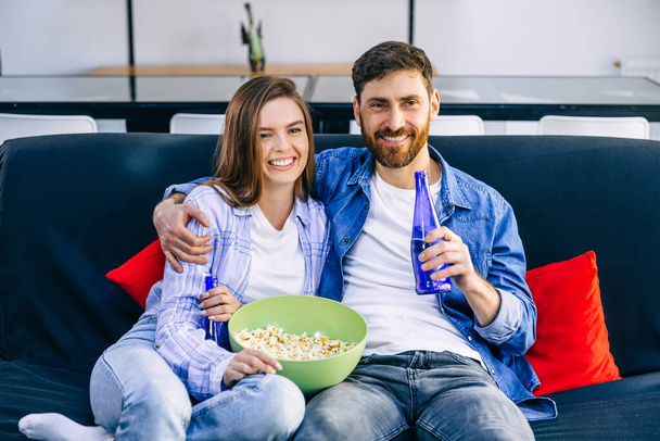 Um jovem casal está assistindo TV no sofá, bebendo cerveja e comendo pipocas. - Foto, Imagem