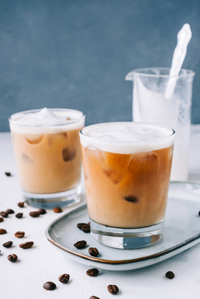 Ledová káva ve sklenici s mléčnou pěnou a kostkami ledu, kávová zrna na bílém stole - Fotografie, Obrázek