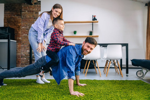 Happy funny family. son playing with father at home, young dad crawling on the floor carrying cute little baby on his back, giving the baby a ride on his back, having fun together. Mom stands, smiles and controls. - Zdjęcie, obraz