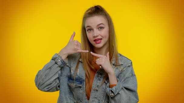Teenager girl in denim jacket looking at camera doing phone gesture like says hey you call me back - Záběry, video