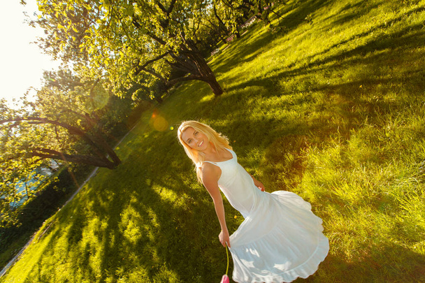 Όμορφη νεαρή λευκή κοπέλα με λευκό φόρεμα γυρίζει στο πάρκο - Φωτογραφία, εικόνα