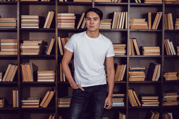 Asian man in white T-shirt stands against the background of bookshelves. Mockup. - Фото, изображение