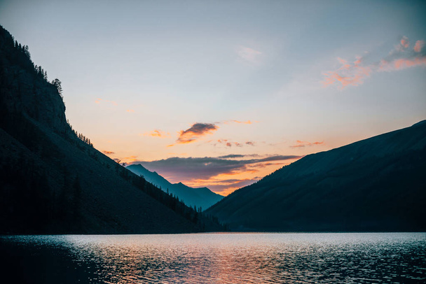Canlı gündoğumu gökyüzü dağ siluetlerinin yakınındaki saf alp gölüne yansıyor. Şafak renklerinde dağ gölünün sakin sularıyla renkli bir manzara. Güneşli bir sabahta su dalgalanması olan güzel bir manzara.. - Fotoğraf, Görsel