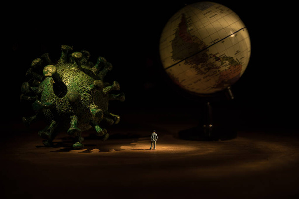 Corona virus global pandemic concept. Miniature man standing near big Corona virus novel in dark. Creative decoration with fog and backlight. Selective focus - Photo, Image