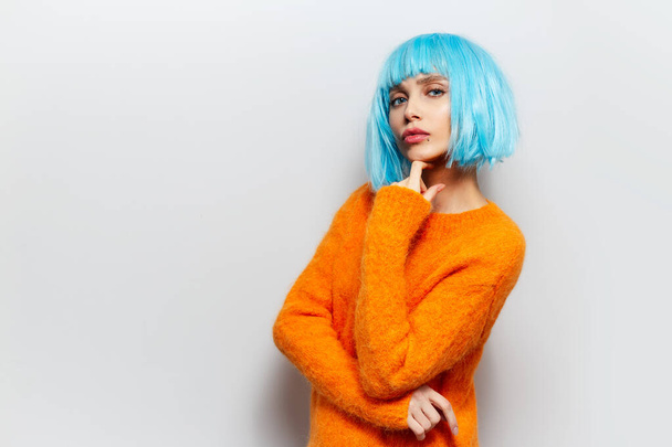 Beyaz arka planda, mavi peruk ve turuncu kazak giyen güzel bir kızın stüdyo portresi.. - Fotoğraf, Görsel
