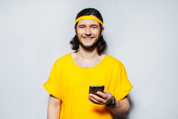 Portrét mladého usmívajícího se hipstera držícího smartphone v ruce, oblečeného ve žluté košili na pozadí šedé texturované stěny. - Fotografie, Obrázek