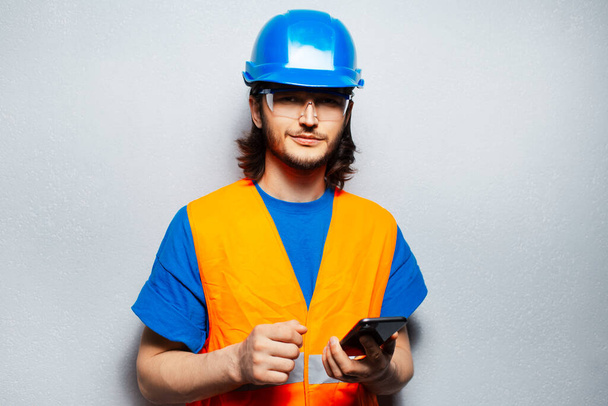 Студийный портрет молодого инженера-строителя, использующего смартфон, смотрящего в камеру, носящего защитное оборудование на фоне серой текстурированной стены. - Фото, изображение