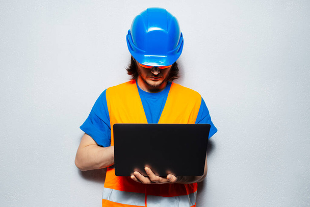 Studio portrait of young man construction worker engineer wearing safety equipment, using laptop on background of grey wall. - Photo, Image