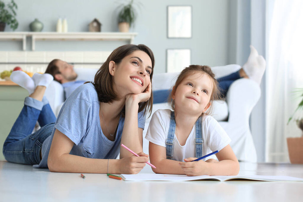 Happy mother smilling daughter lying on warm floor enjoying creative activity, drawing pencils coloring pictures in albums, father resting on couch, family spend free time together - Фото, изображение