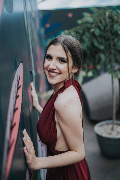Una hermosa morena sonriente con un elegante vestido rojo posando junto a la pared decorada de un café al aire libre - Foto, imagen