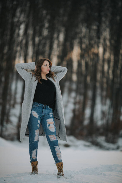 A full body shot of a brunette woman posing outside in winter wearing along gray coat and ripped jeans - Photo, image